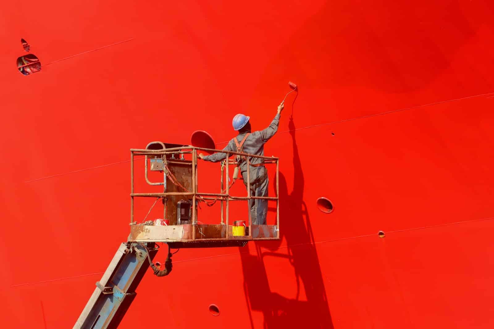 Worker painting ship hull on sherry picker using paint roller
