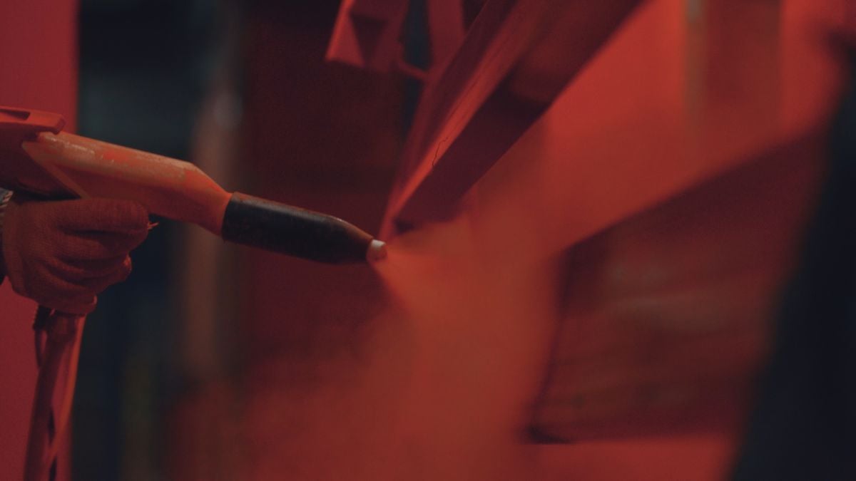 Worker's gloved hand operates an electrostatic spray gun, applying a vibrant red powder coating