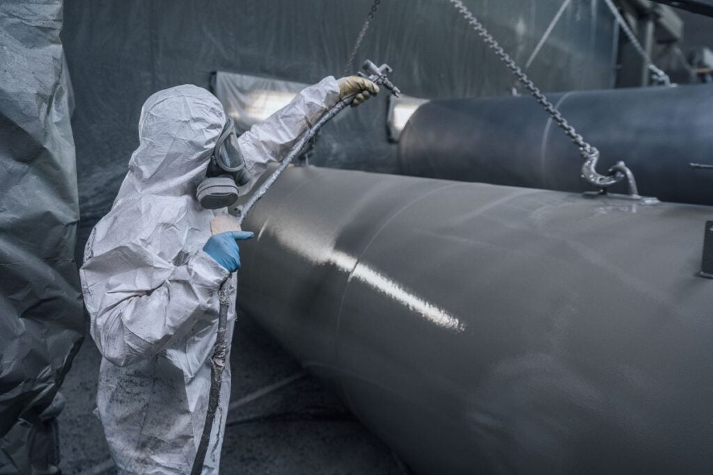 worker wearing protective gear and mask spray painting tanks