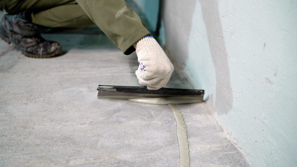 waterproofing commercial building Closeup of worker hand in glove using trowel to spread gray sealant along concrete floor edge, renovation and repair concept