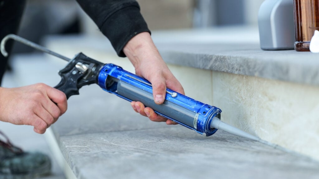 A person uses a blue caulk gun to apply a bead of sealant along the edge of outdoor stone steps, ensuring a watertight seal