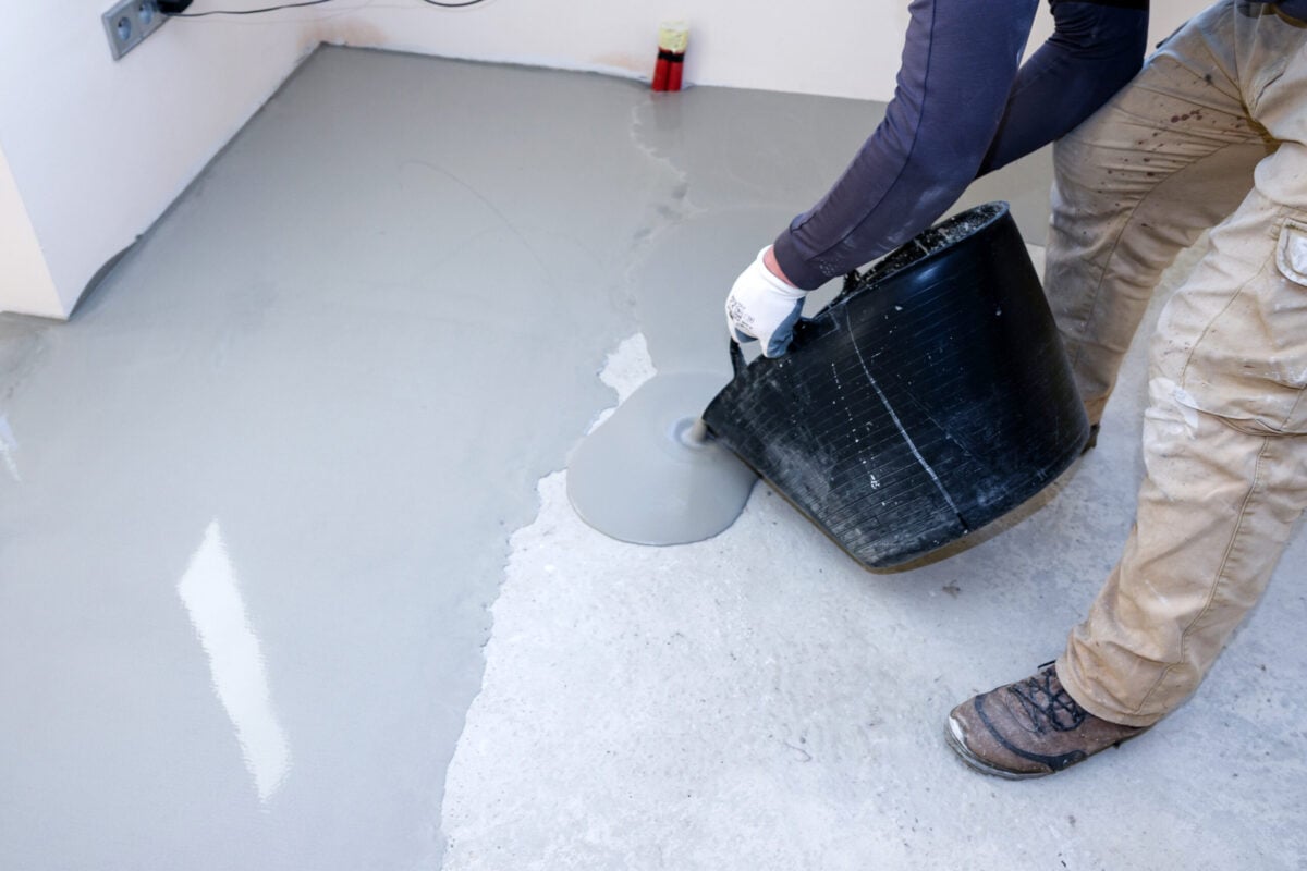 self-leveling underlayment A worker is pouring a cement mixture from a bucket onto a concrete self-leveling floor in a construction area. The activity takes place inside a building during the day.