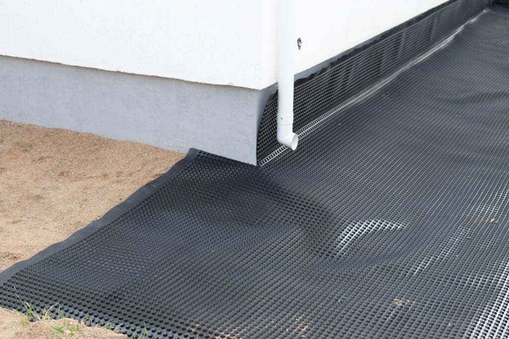 Laying a black waterproofing membrane around the foundation of a house. Construction of the blind area of the house. Reliable protection of the foundation from moisture. White pipe for draining sediment from the roof of the house. Horizontal photo