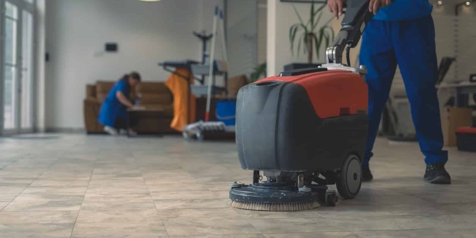 Cleaner cleans hard floor with scrubber machine while other cleaner cleans in the background